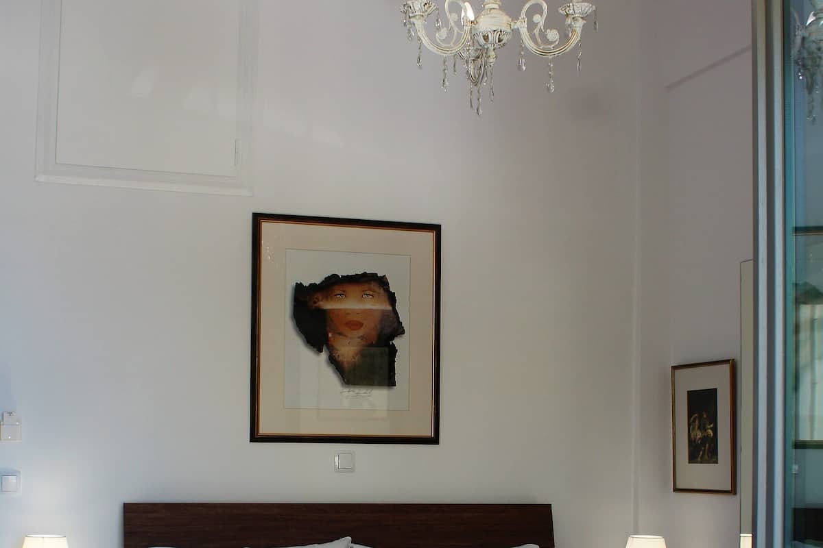 Uma cama bem arrumada com lençóis brancos e colcha laranja, um candelabro e quadros nas paredes.