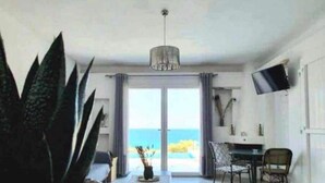 Ein modernes Wohnzimmer mit einer großen Topfpflanze, einer Couch, einem Essbereich mit Stühlen, einem an der Wand befestigten Fernseher und Meerblick durch große Fenster.