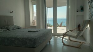 Ein Schlafzimmer mit Bett, Stuhl, Fenster mit Meerblick und einer Zimmerpflanze.