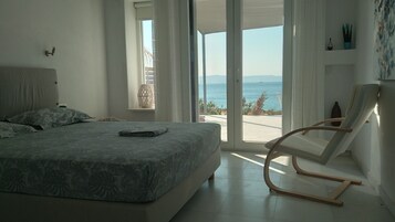 A bedroom with a bed, a chair, a window with a view of the sea, and a potted plant.