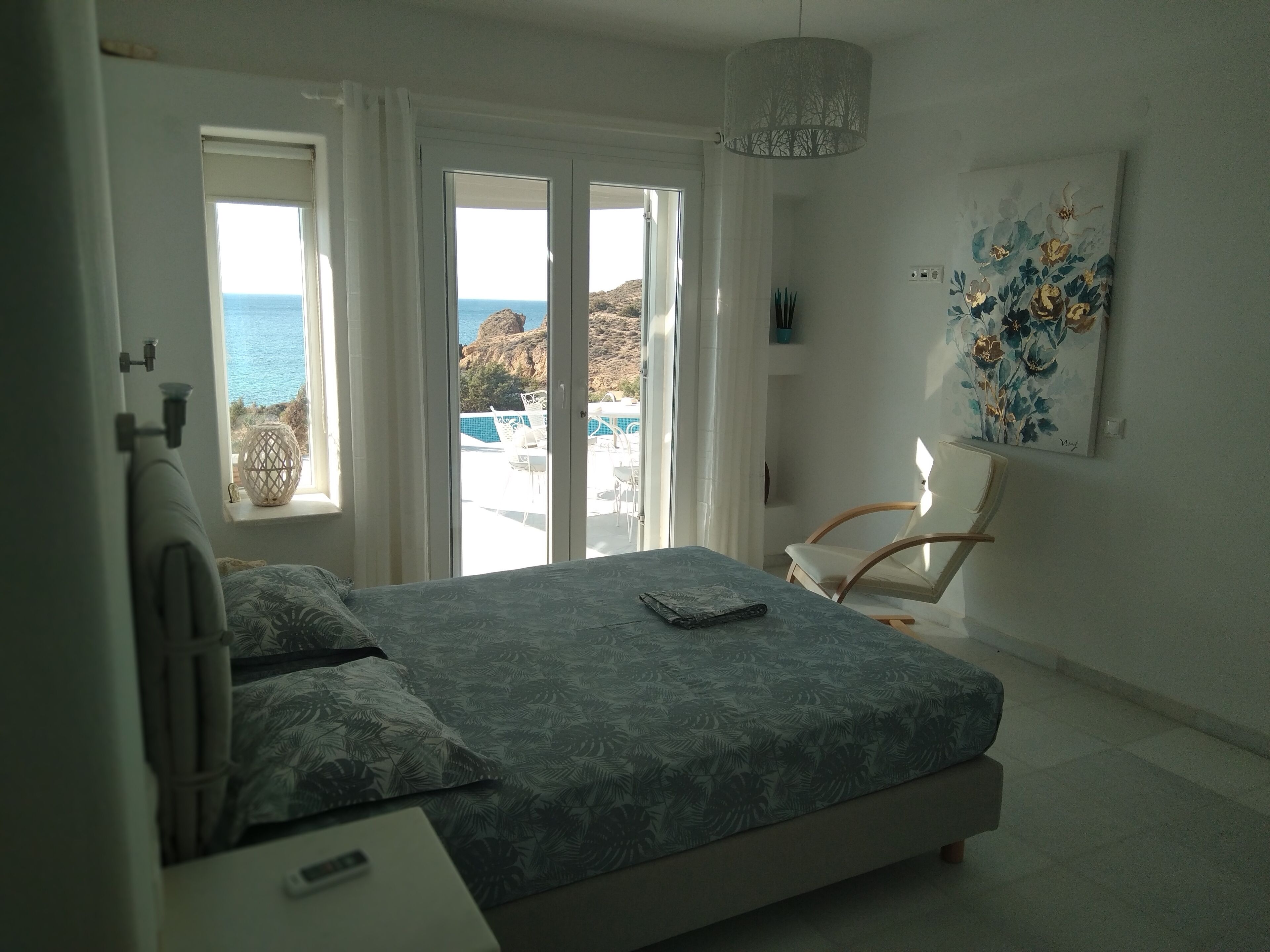 A bedroom with a bed, a chair, a painting, and a view of the sea through a door.