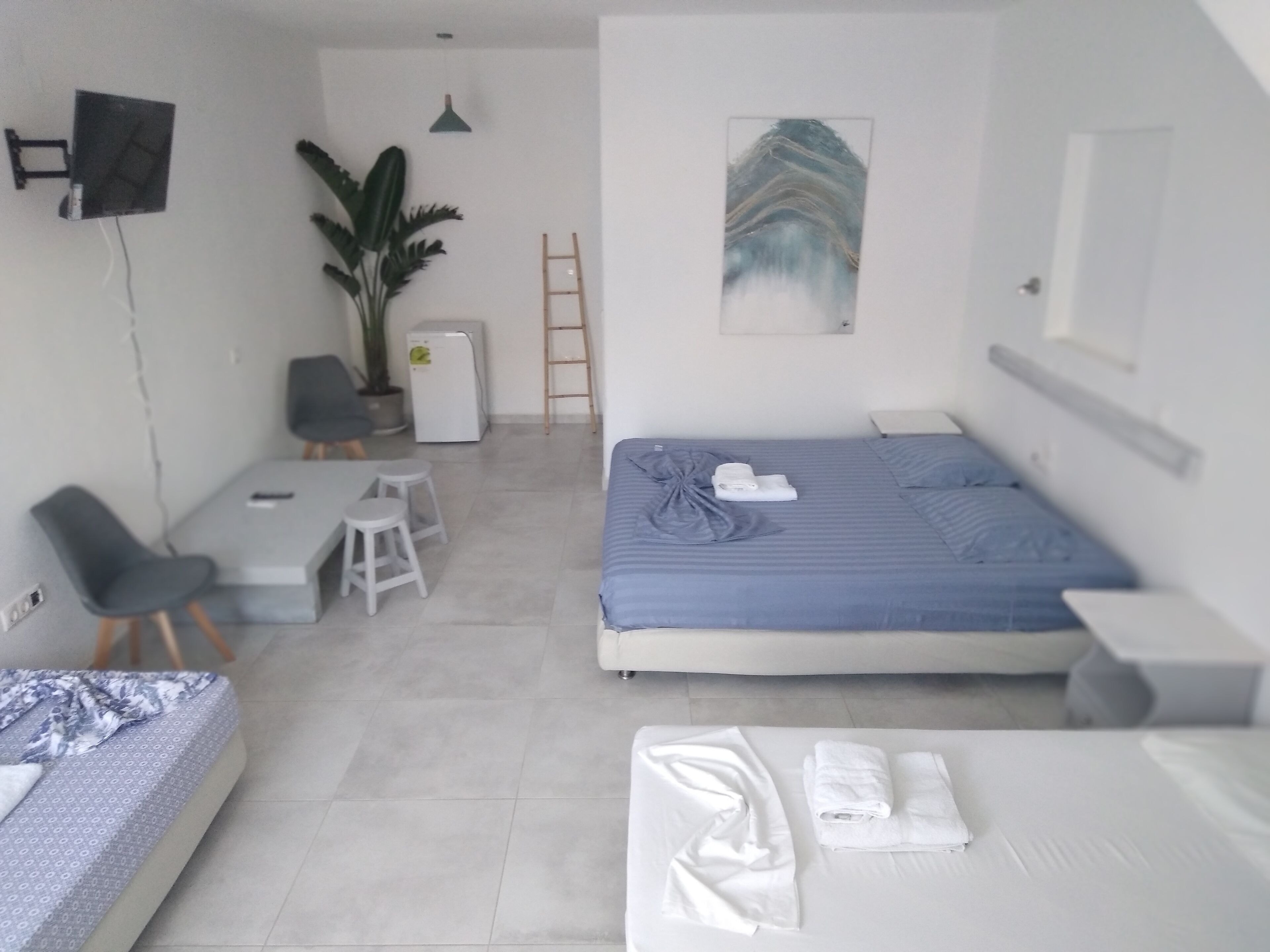 A modern, well-lit room with a bed, a desk, and a chair. There is a plant, a ladder, and a painting on the wall.