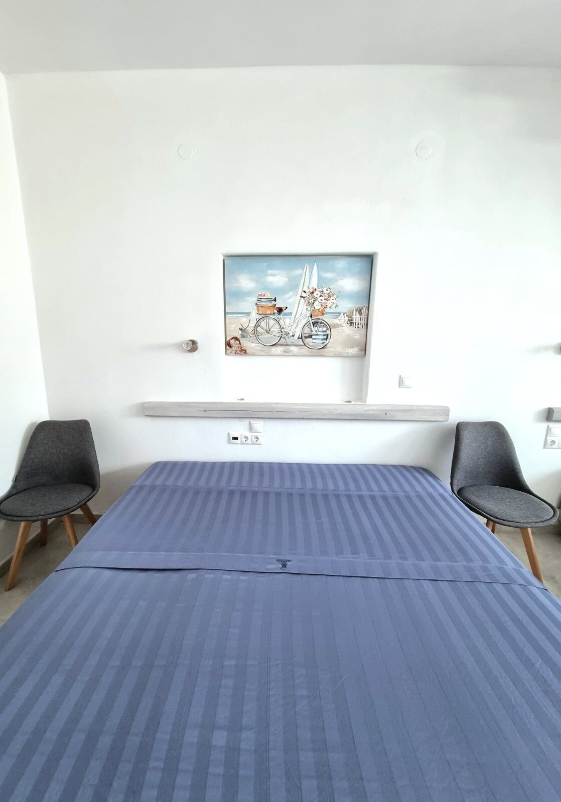 A room with a bed, two chairs, a painting of a beach scene, and a small shelf with a heater.