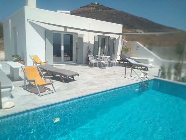 A modern outdoor pool area with lounge chairs and a table, surrounded by a white building with glass doors. - Glymar (Paros)