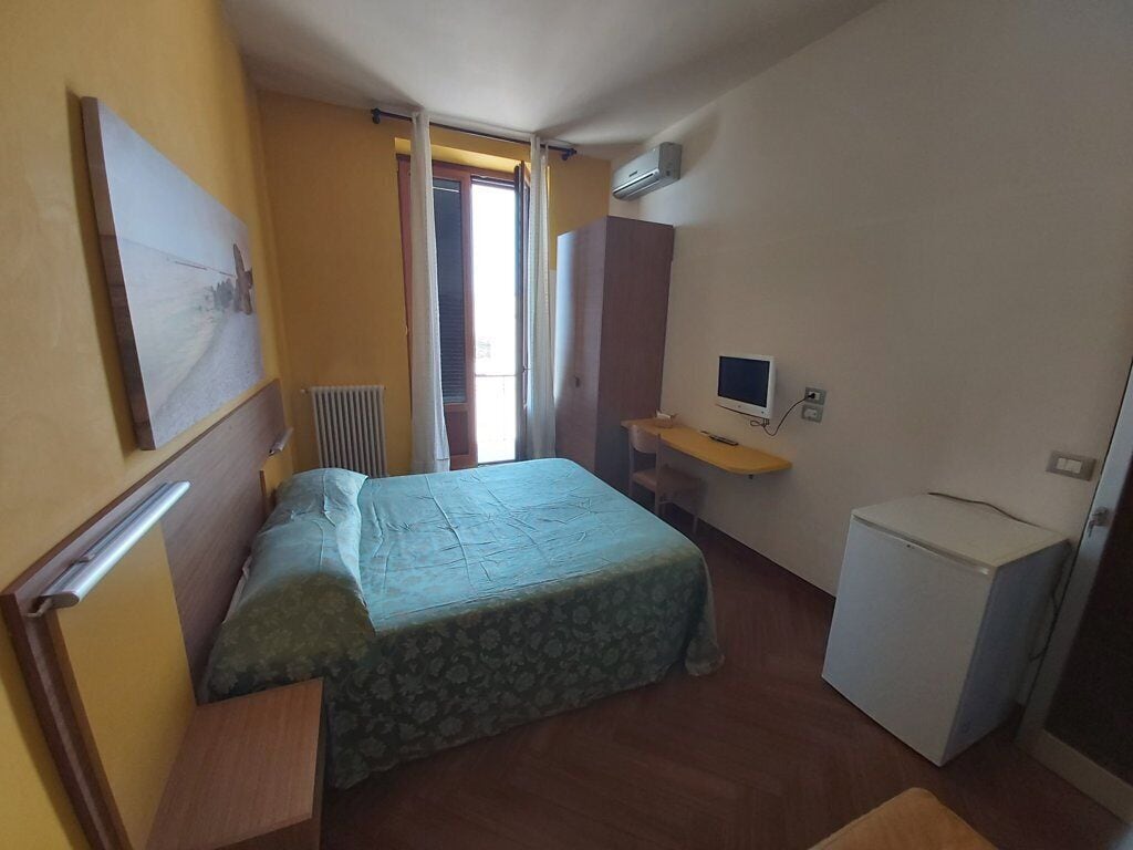 Een kleine hotelkamer met een bed, een bureau, een stoel, een radiator en een koelkast.