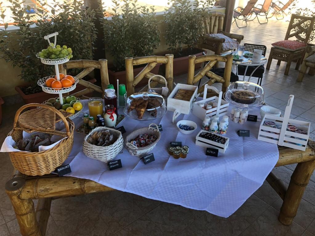 Une table de buffet proposant une variété de plats et de boissons, notamment des fruits, des pâtisseries et des condiments, installée à l’extérieur avec des chaises et des tables.