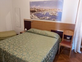 Habitación de hotel con cama, una silla, una mesita de noche y un cuadro colgado en la pared que muestra un puerto deportivo.