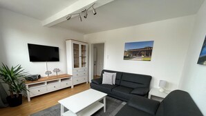 Una sala de estar moderna con un televisor de pantalla plana, un sofá negro, una mesa de centro blanca y un mueble con puertas de vidrio.