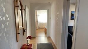 Un pasillo con suelo de madera, una pequeña mesa de madera con un jarrón de flores, un tapete gris y un espejo en la pared izquierda.