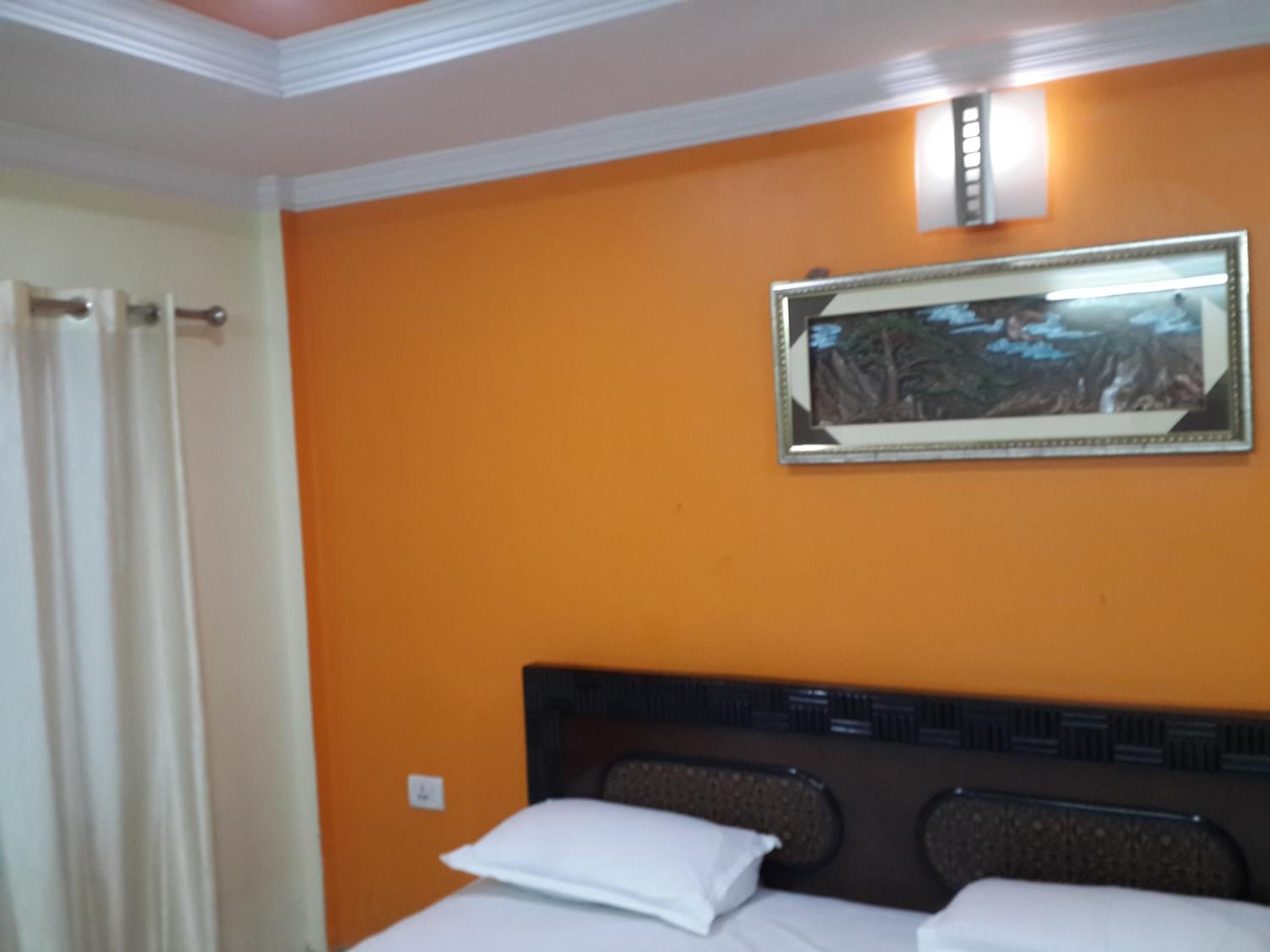 A hotel room with an orange wall, a framed picture, a bed with white bedding, and a white curtain.