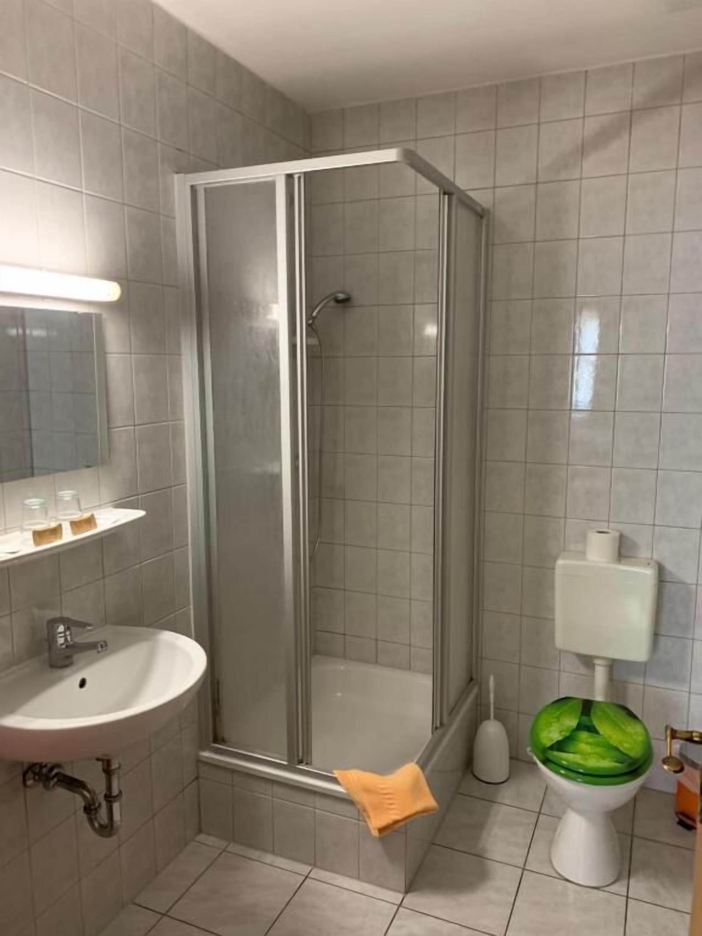 Un baño con ducha de mampara de vidrio, un lavamanos con espejo y un inodoro con asiento verde.