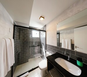 Un baño moderno con ducha revestida de mosaicos blanco y negro, inodoro blanco, lavabo negro y un espejo grande.