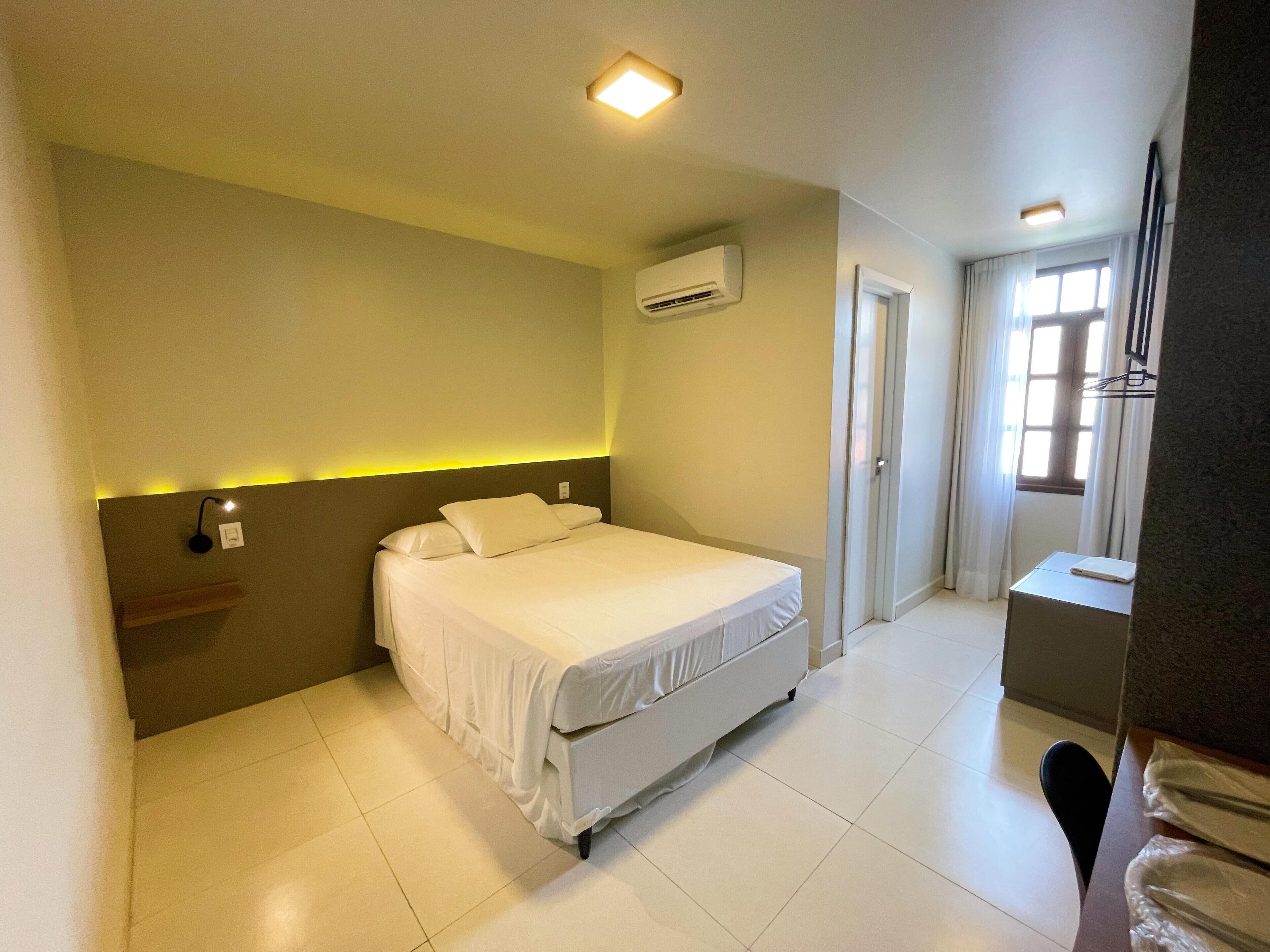 A hotel room with a bed, a bedside table, an air conditioning unit, a door, and a window.