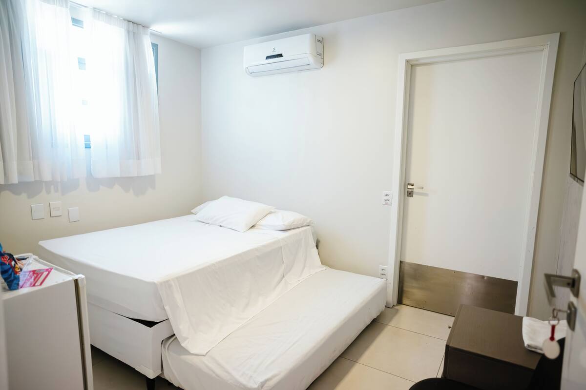 Una habitación pequeña y limpia con una cama individual, un escritorio, una silla, una puerta y un equipo de aire acondicionado en la pared.