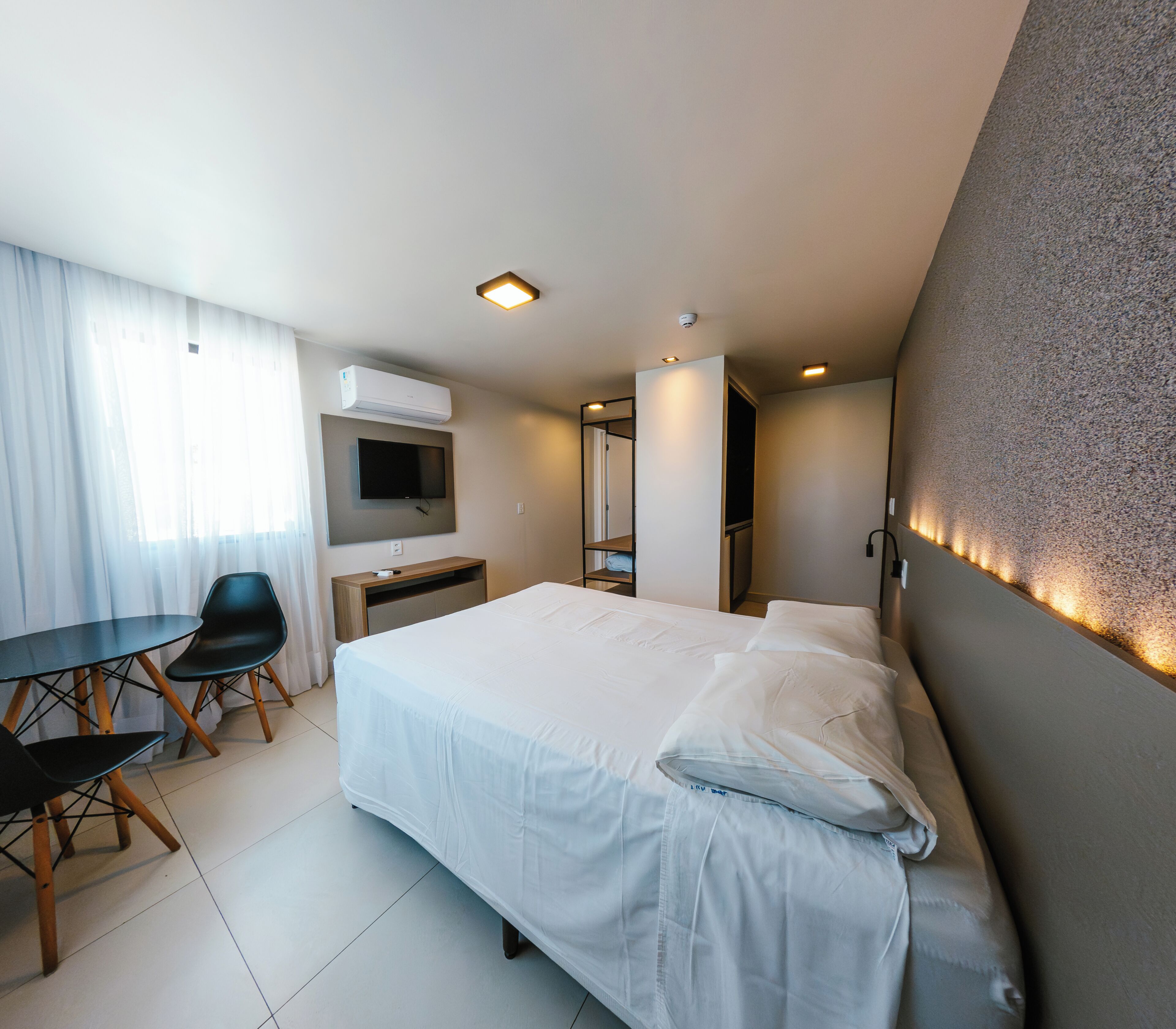 A hotel room with a bed, a desk, a chair, a television, and an air conditioning unit.