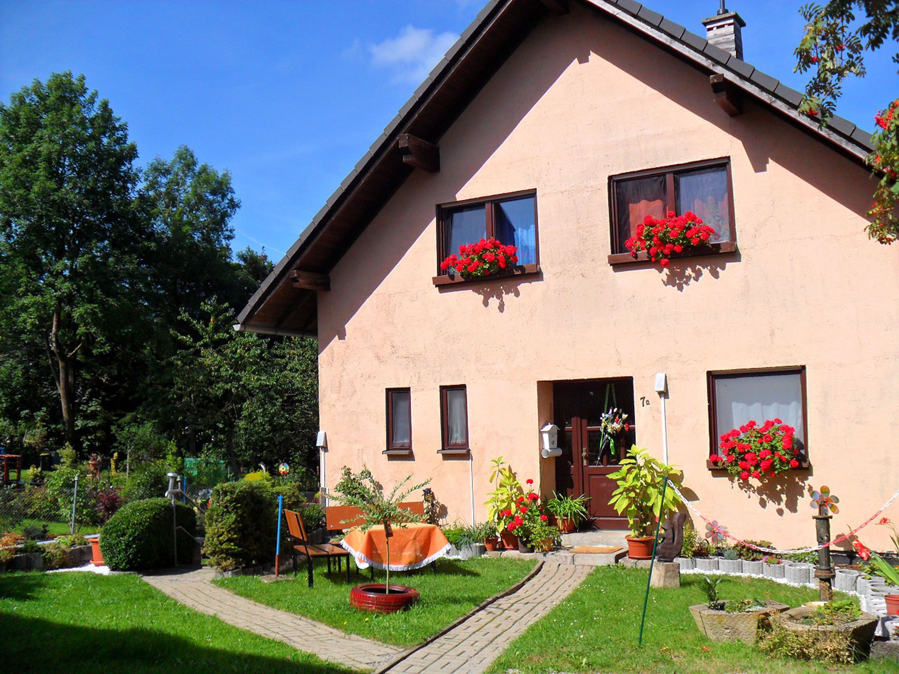 A two-story house with a sloped roof, multiple windows, and a front door. There are flower boxes with red flowers on the windowsills, and a paved pathway leading to the entrance.