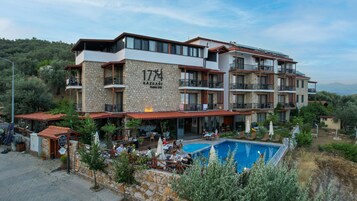 Un hotel de varios pisos con fachada de piedra, balcones, mobiliario exterior y una piscina.