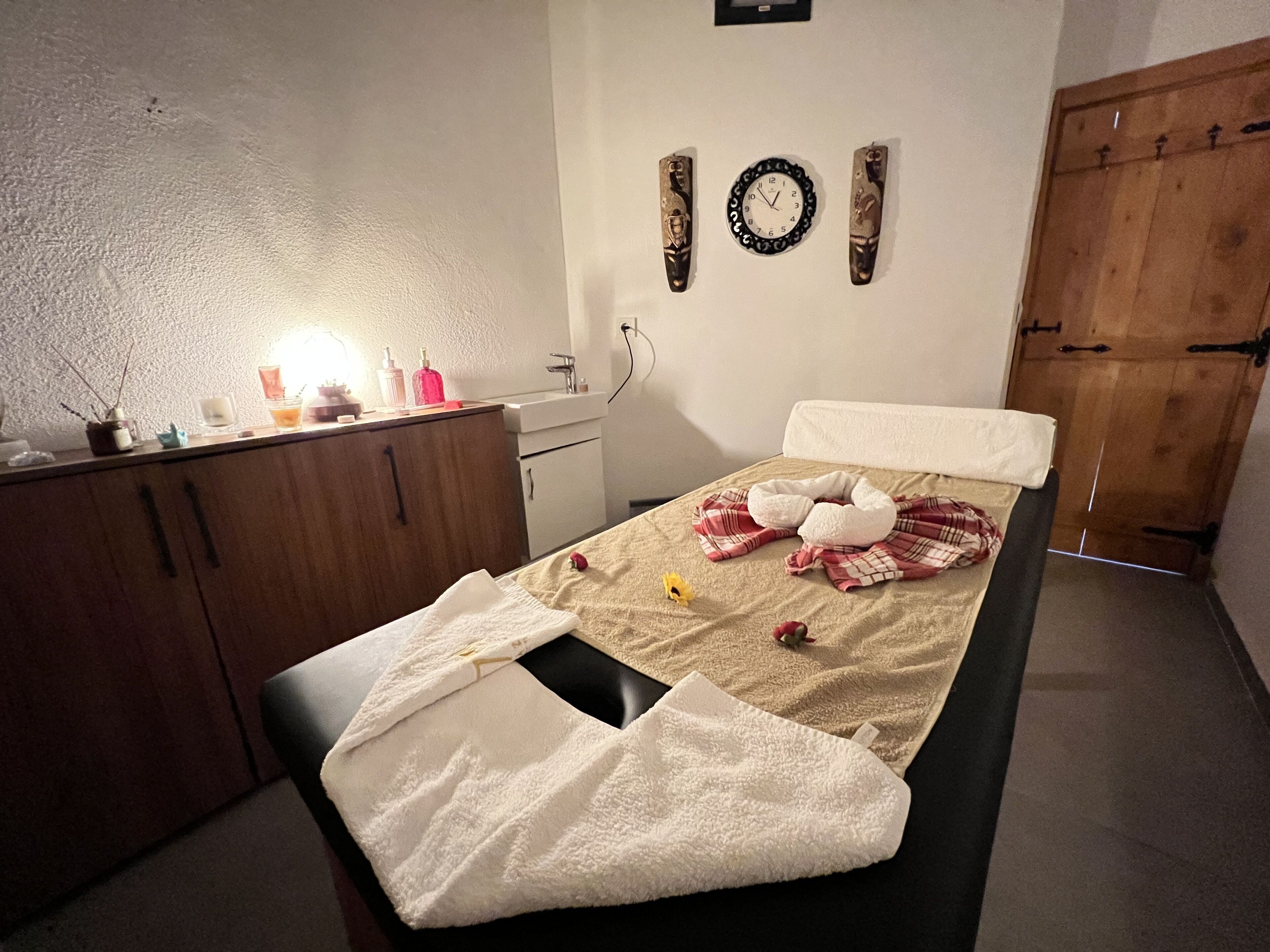 A bedroom with a bed, a wooden wardrobe, a bedside table with toiletries, a wall clock, and a wooden door.