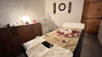 A bedroom with a bed, a wooden wardrobe, a bedside table with toiletries, a wall clock, and a wooden door.