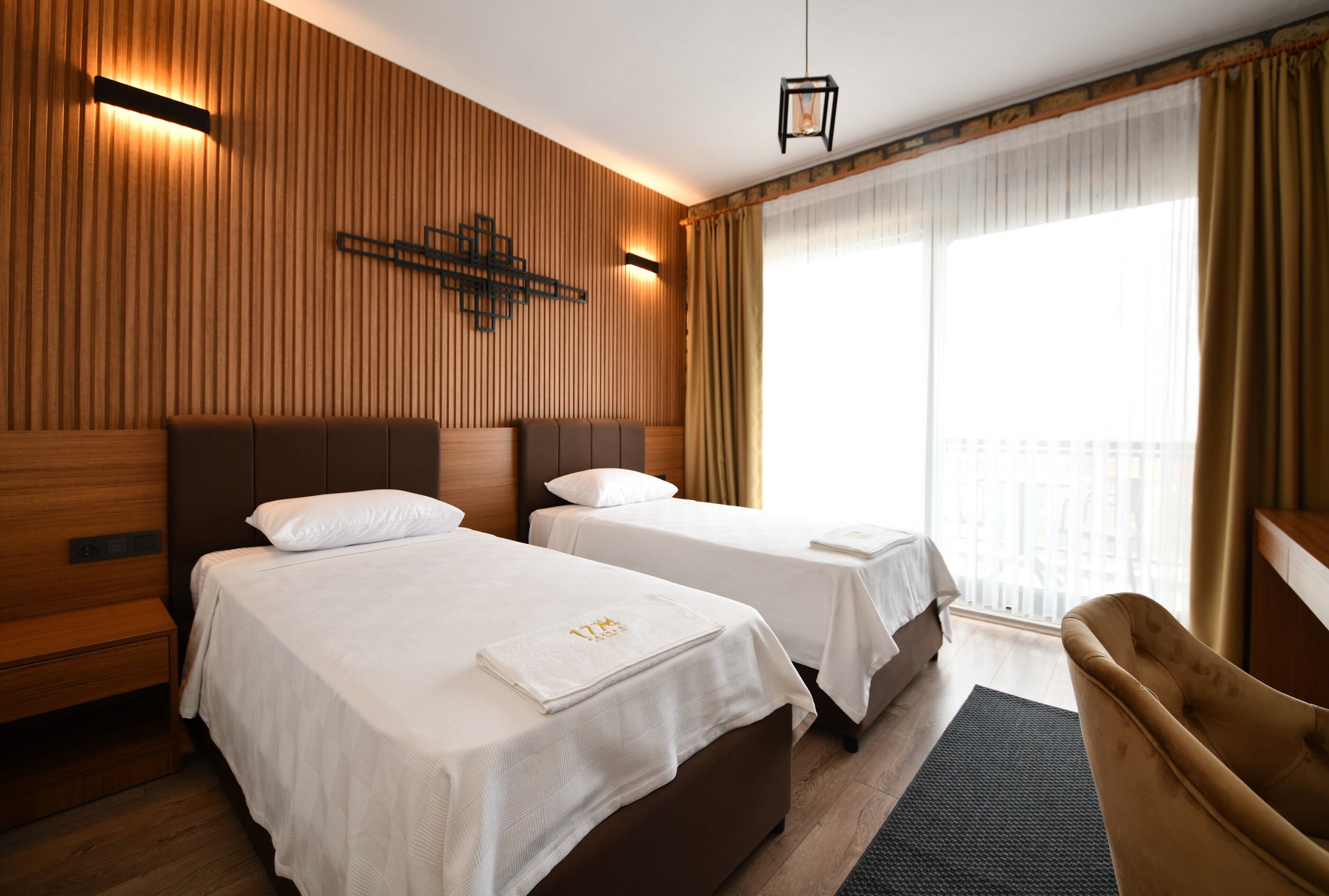 A hotel room with two beds, a wooden wall paneling, a hanging light fixture, and a view of the outdoors through large windows.