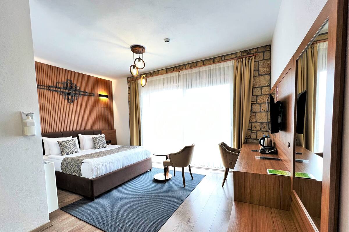 Ein Hotelzimmer mit Bett, Schreibtisch, Stühlen, Fernseher und einem großen Fenster mit Vorhängen.
