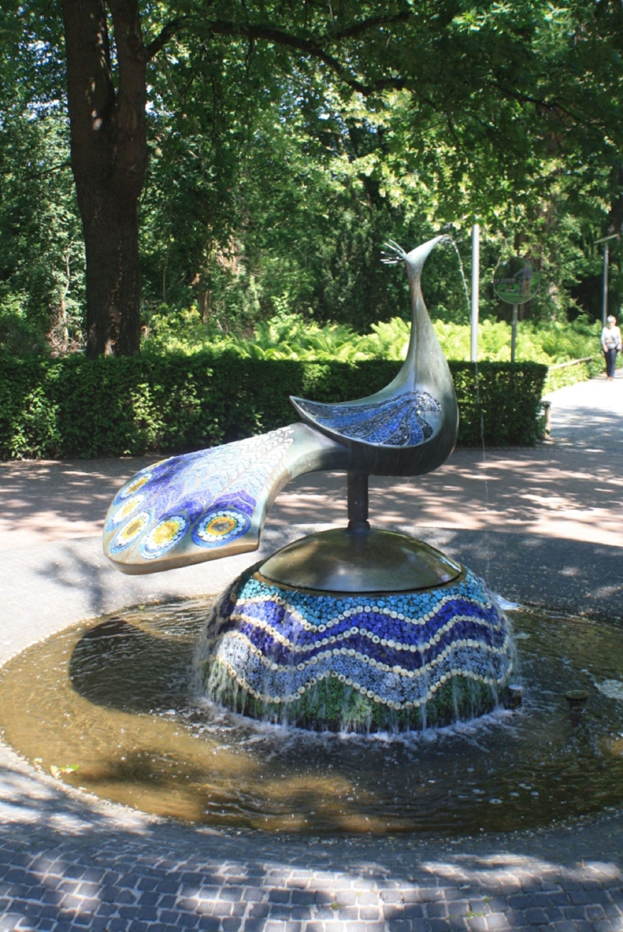 Una fuente con diseño de pavo real, rodeada de follaje y un camino pavimentado.