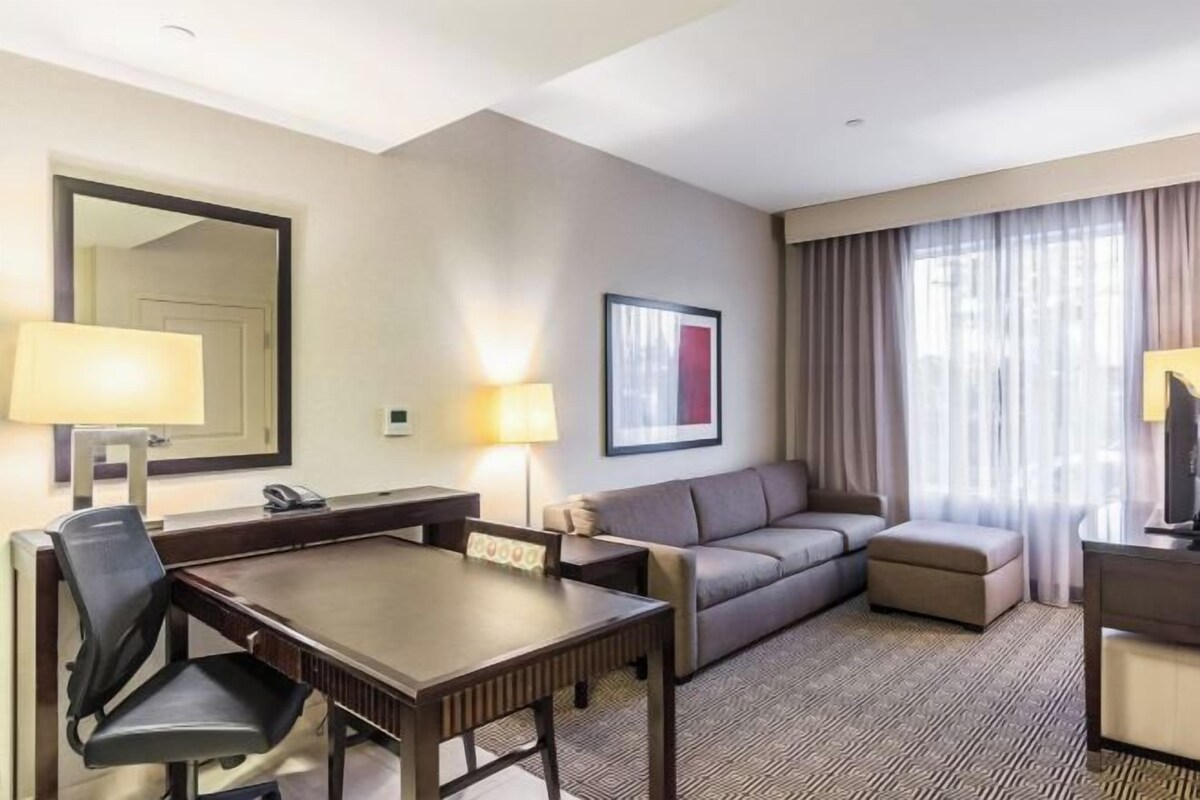 Une chambre d’hôtel équipée d’une table à manger, de deux chaises, d’un canapé, d’un petit pouf, d’une télévision et d’une grande fenêtre avec des rideaux.