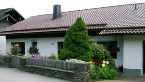Uma casa com telhado de cerâmica, parede de pedra e diversos flores e plantas em frente.