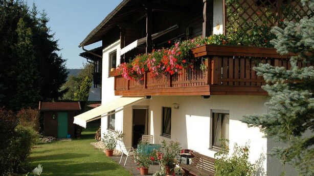 Uma casa de dois andares com uma varanda decorada com flores, um pequeno jardim e um deck de madeira.