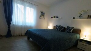 A bedroom with a bed, a nightstand, a lamp, a shelf with books, and a window with curtains. - Ferienwohnung Auszeit (Hofheim in Unterfranken)