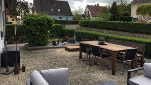 A patio area with a wooden table and chairs, a grey sofa, and a small garden with a hedge and a bird feeder. - Ferienwohnung Auszeit (Hofheim in Unterfranken)