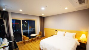 Habitación de hotel con una cama grande, un escritorio, una silla y una mesita. Hay una ventana con cortinas y se ve una vista nocturna de la ciudad desde el exterior.