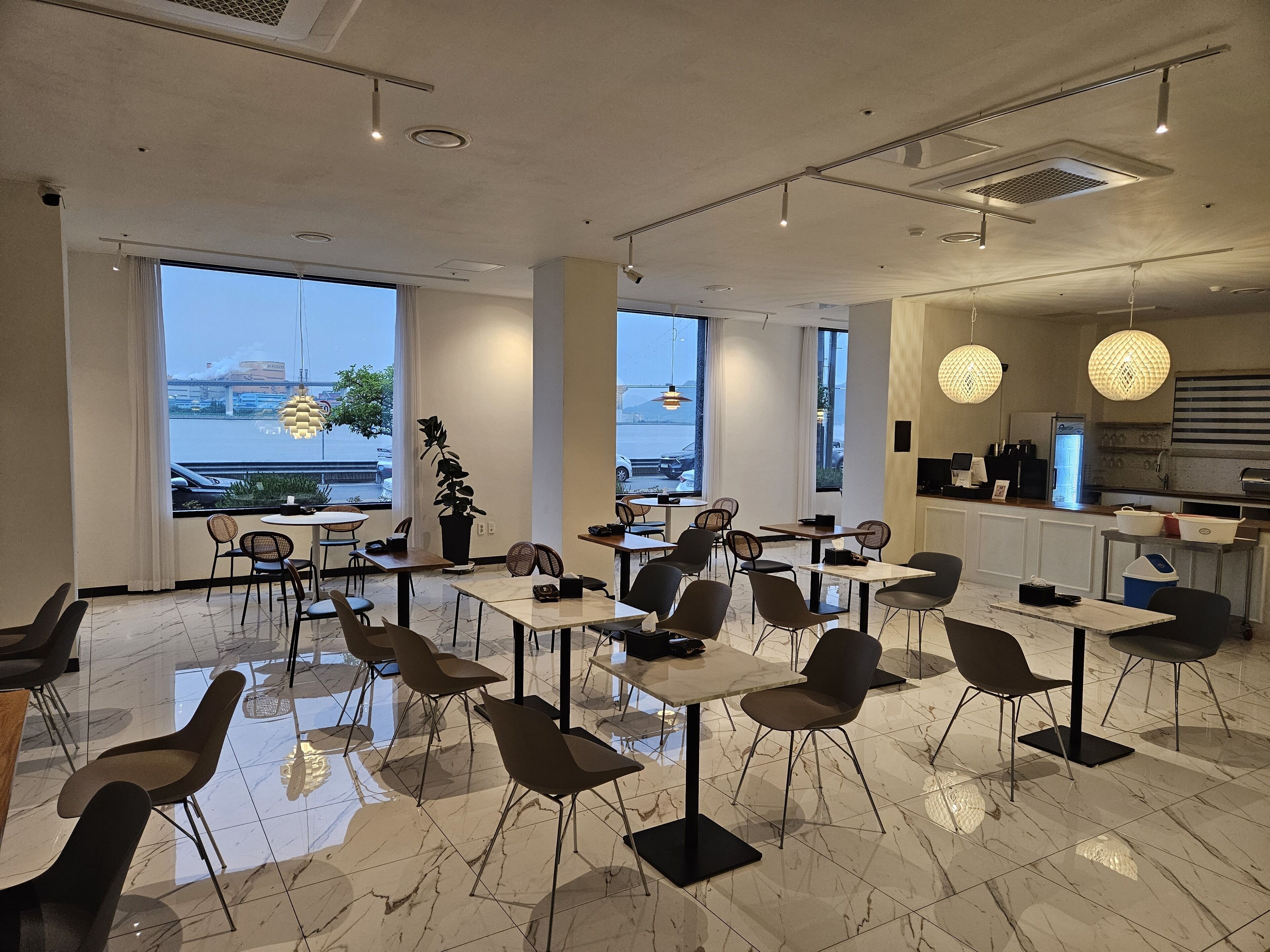 A modern dining area with marble flooring, multiple tables and chairs, large windows with a view of the water, and pendant lighting.
