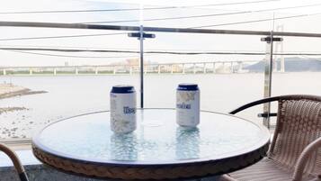 Mesa de vidro redonda com duas latas de cerveja, duas cadeiras de vime e vista para um rio e uma ponte.