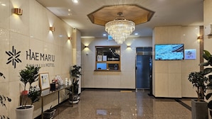 Zona de recepción de hotel con suelo de mármol pulido, mostrador de recepción, candelabro y una pantalla de televisión que muestra un paisaje nevado.