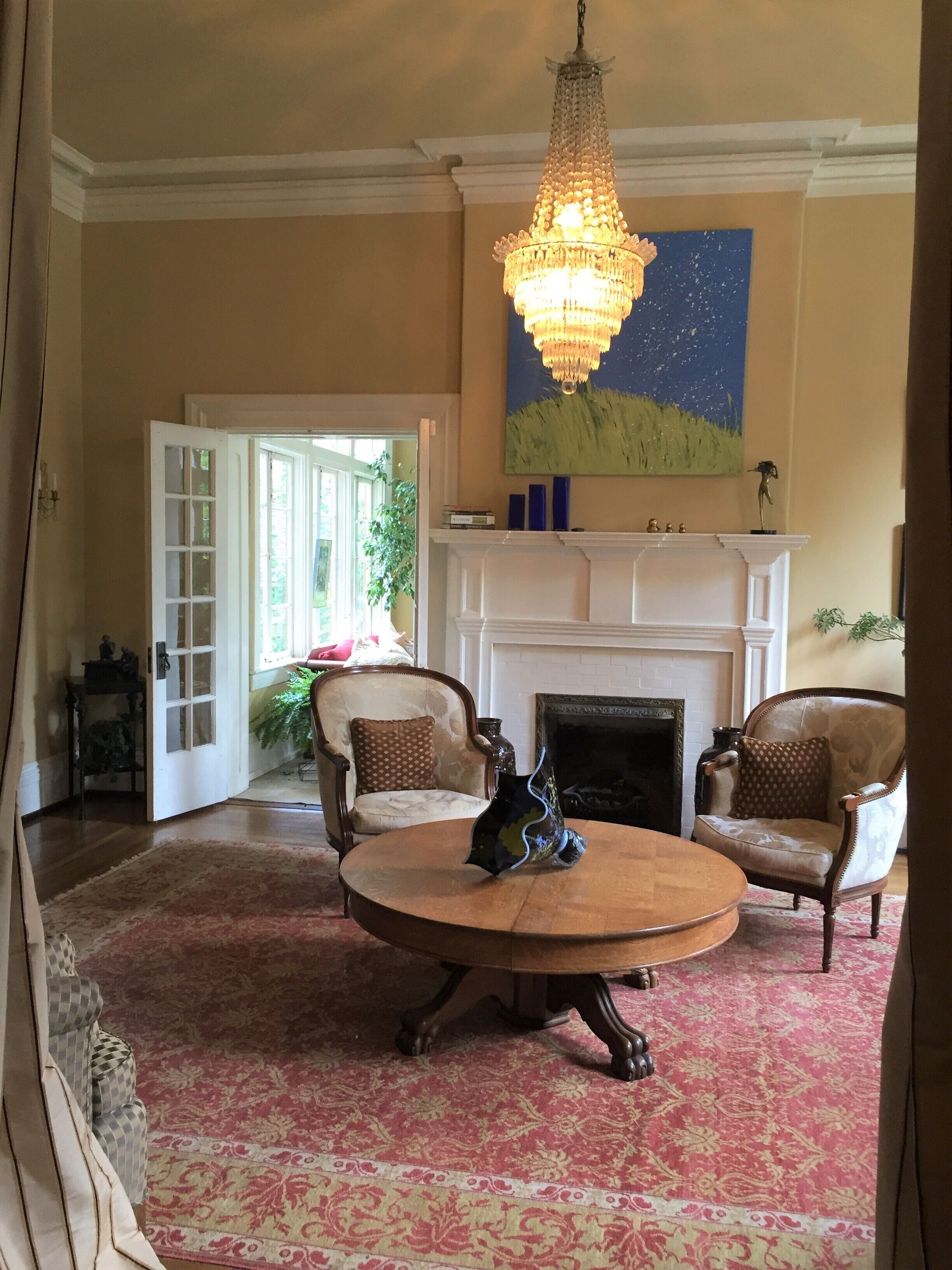 A room with a round wooden table, two armchairs, a white fireplace, a chandelier, and a painting of a landscape.