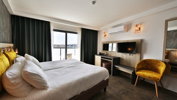 A hotel room with a large bed, a desk, a chair, a TV, and a view of the city. - Ivy Hotel - Adults Only (St. Julian's)