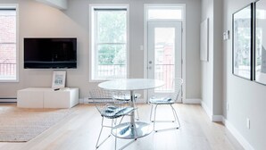 Un espace repas moderne avec une table ronde et des chaises en métal, un téléviseur à écran plat fixé au mur et de grandes fenêtres laissant entrer la lumière naturelle.