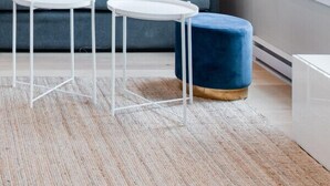 Un salon moderne avec un canapé gris, deux petites tables d’appoint blanches, un pouf bleu et une lampe sur pied.