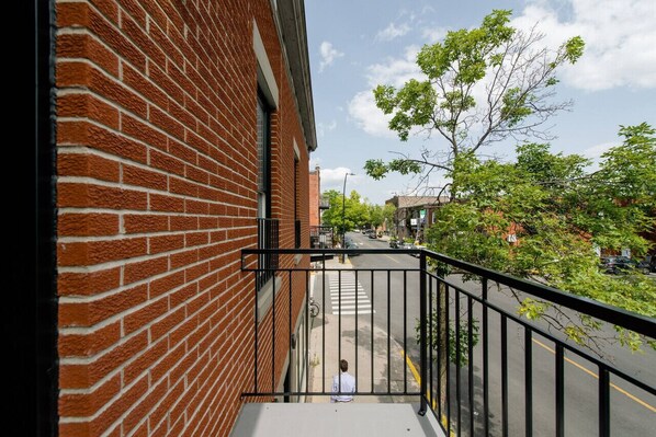 A view from a balcony overlooking a street with a brick building on the left and a tree-lined street.