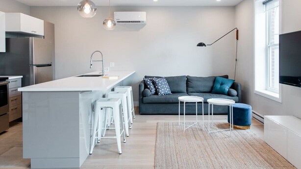 Une cuisine moderne avec un îlot central, des tabourets de bar, un canapé gris et un téléviseur.