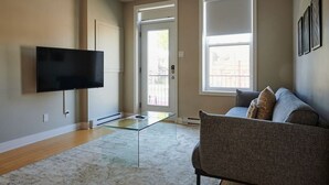 A living room with a flat-screen TV, a glass coffee table, a grey sofa, and a large window with blinds.