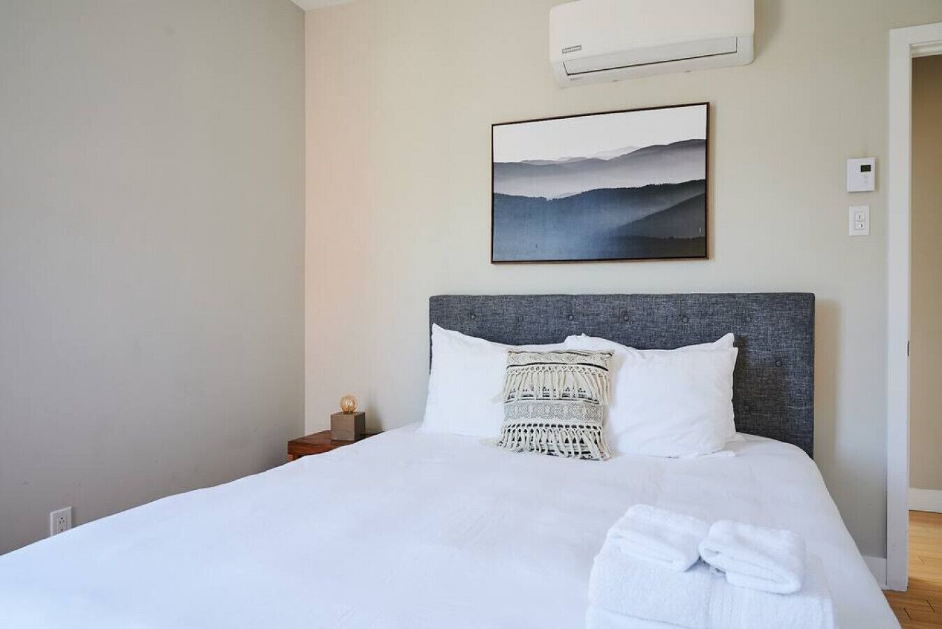 A neatly made bed with white linens and a decorative pillow, a wall-mounted air conditioner, and a framed picture above the bed.