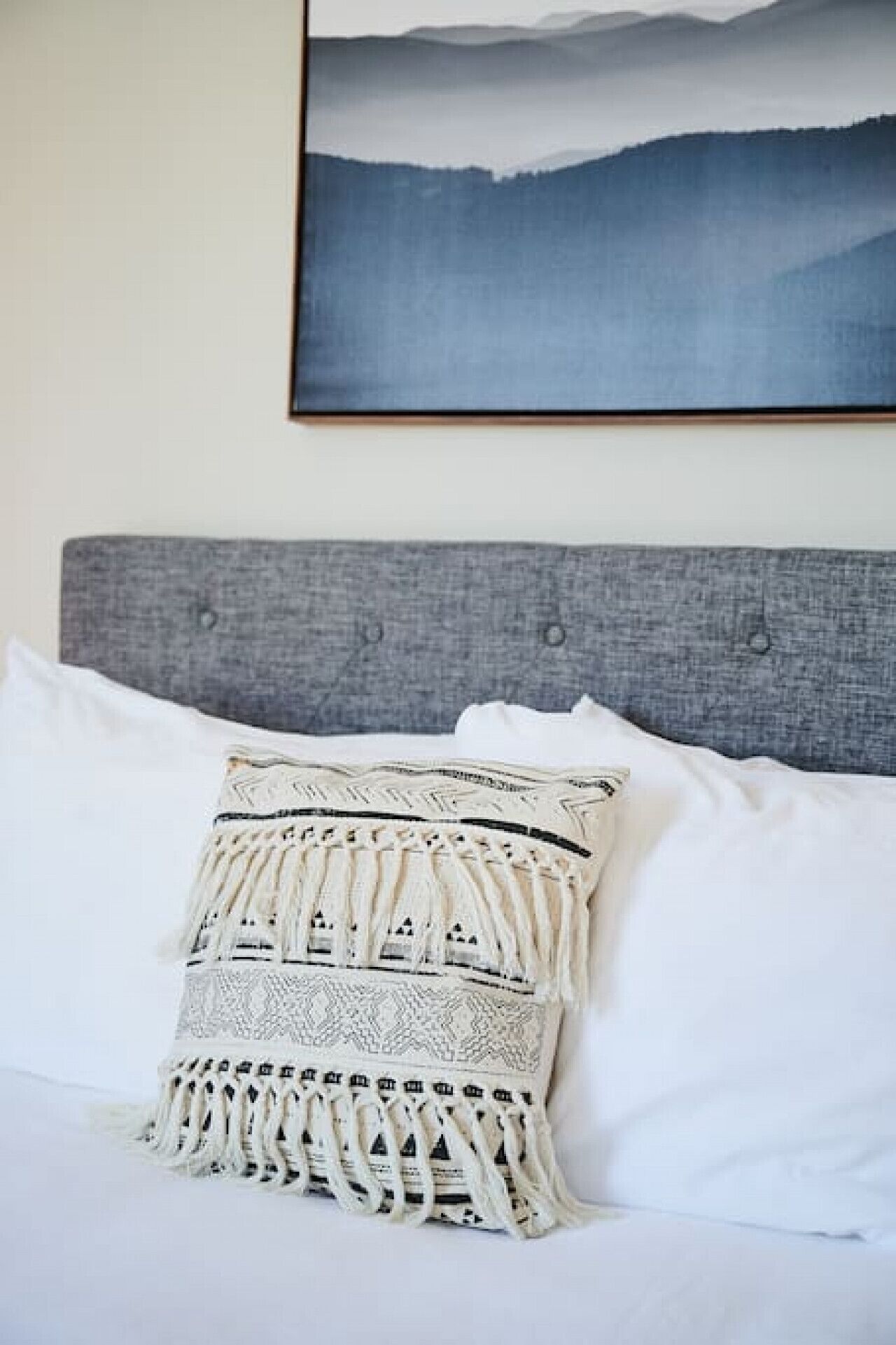 A bed with a grey headboard, white pillows, and a decorative throw pillow with intricate patterns.