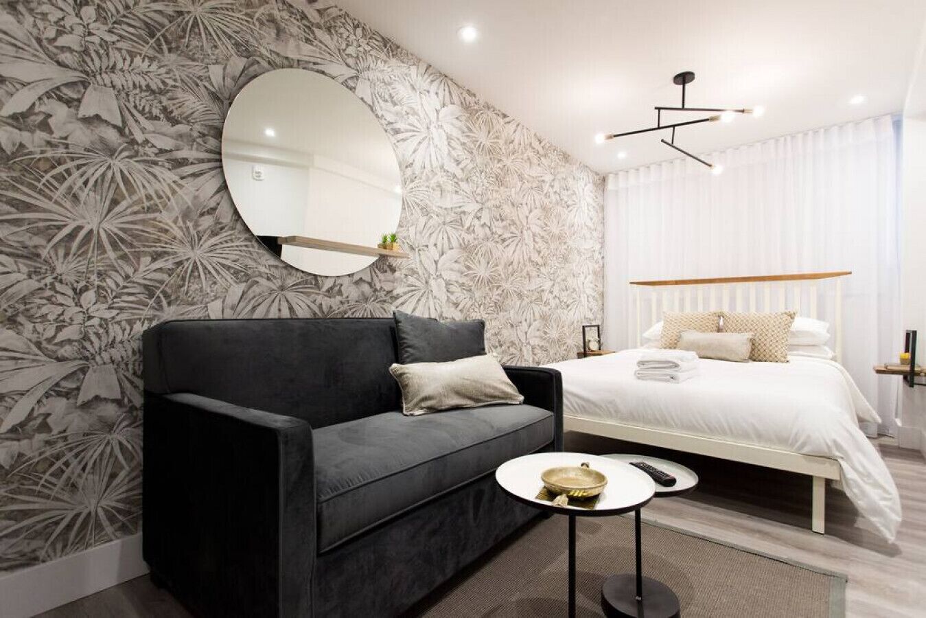 Un dormitorio moderno con un sofá oscuro, una cama con ropa de cama blanca, un espejo redondo y una mesita con un cuenco y un control remoto.