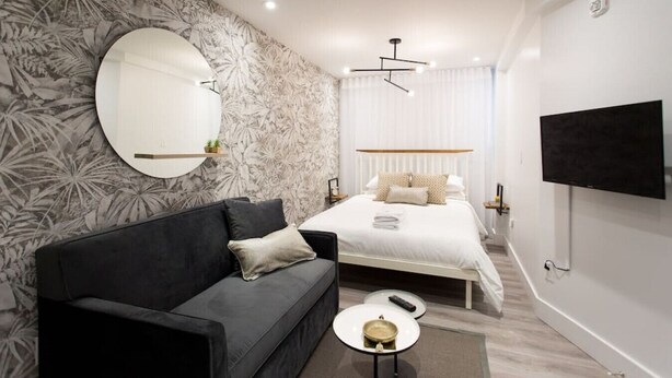 A modern bedroom with a bed, a sofa, a round mirror, a flat-screen TV, and a small table with a remote.