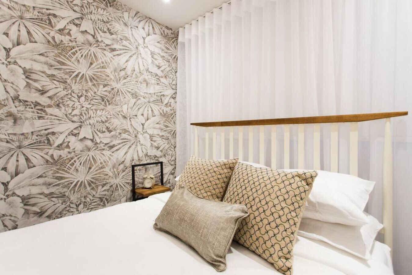 Un dormitorio con cama, almohadas, mesita de noche, reloj y un papel tapiz floral.