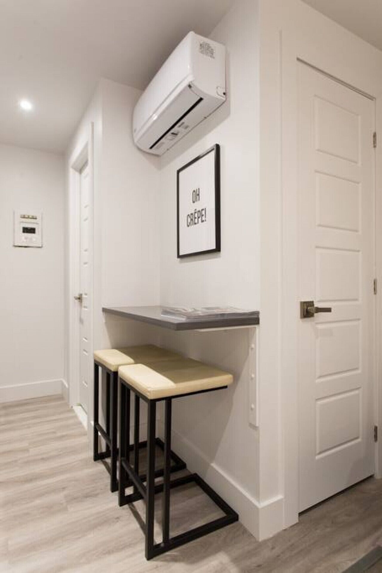 Um corredor com porta branca, um pequeno bar com banquetas, unidade de ar condicionado e uma placa emoldurada na parede.