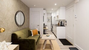 Un espacio habitable compacto con sofá, una mesita, una zona de cocina y una puerta que da a otra habitación.