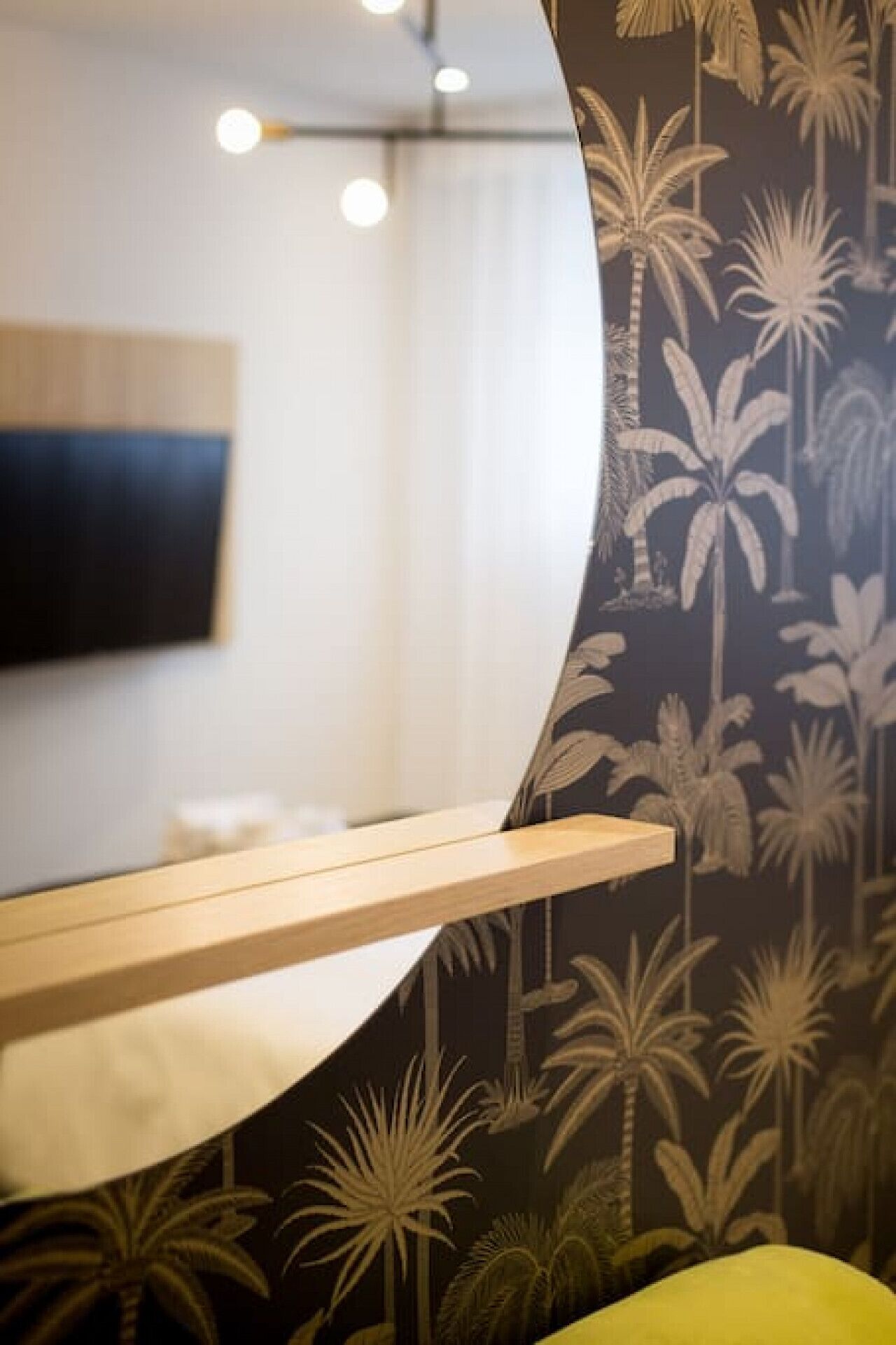 A bathroom with a mirror, a wooden shelf, and a wall with palm tree wallpaper.